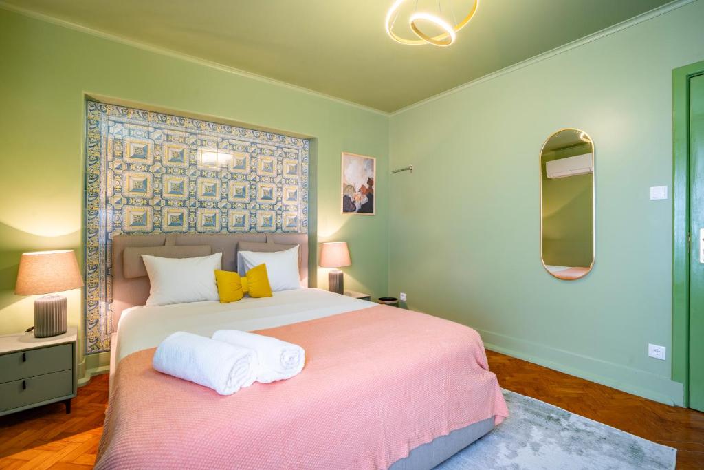 Bedroom with double bed, yellow pillows, pink blanket, patterned wall tiles, lamps, and an oval mirror.