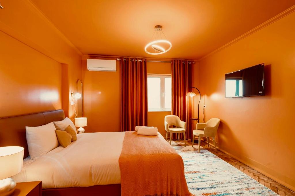Orange-themed bedroom with bed, pillows, window, curtains, two chairs, floor lamp, and wall-mounted TV.