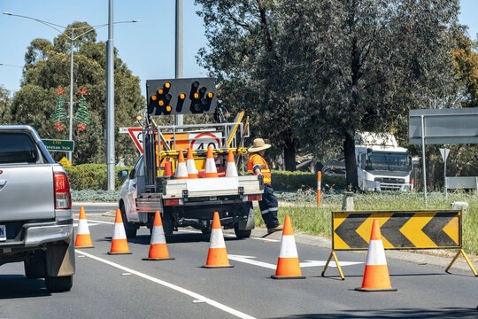 Road maintenance worker wearing high-visibility clothing next to a truck with traffic cones and warning signs on a highway lane.