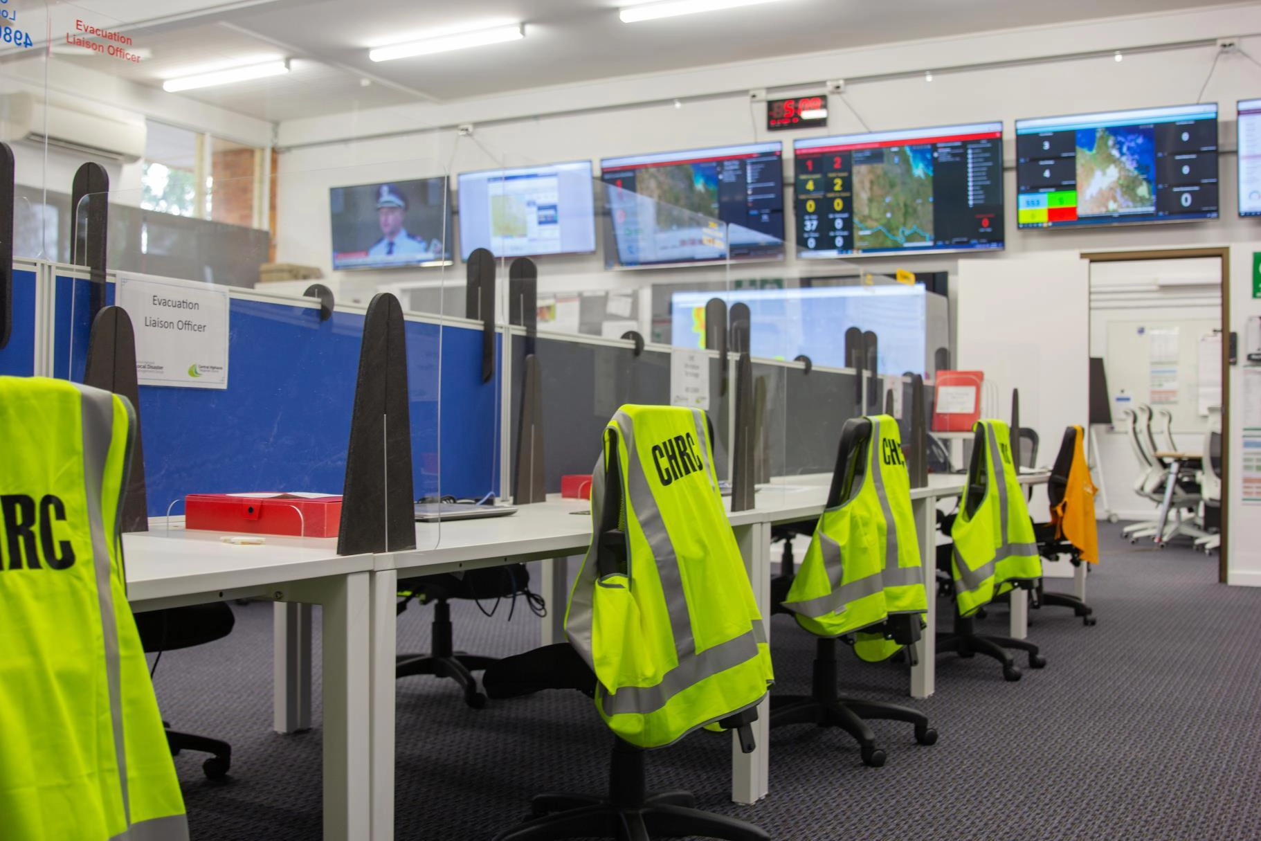 Empty emergency operations center desks with fluorescent yellow CHRC safety vests on chairs and multiple large screens displaying maps and data on the wall.