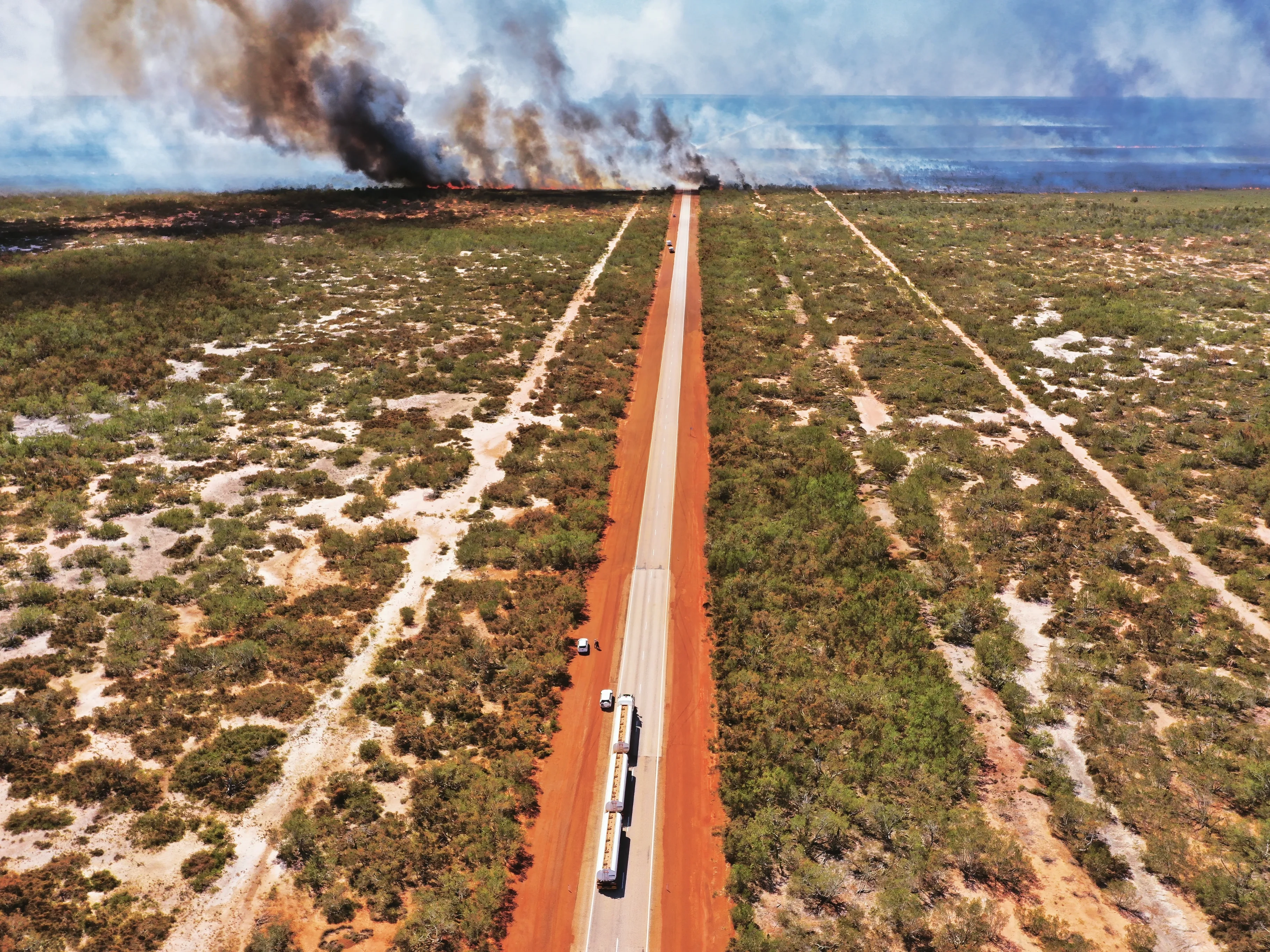 Aerial view of a long straight road through dry bushland with smoke and fire burning in the distance.