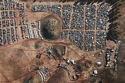 Aerial view of a large encampment with numerous tents and vehicles arranged on dry, barren land.