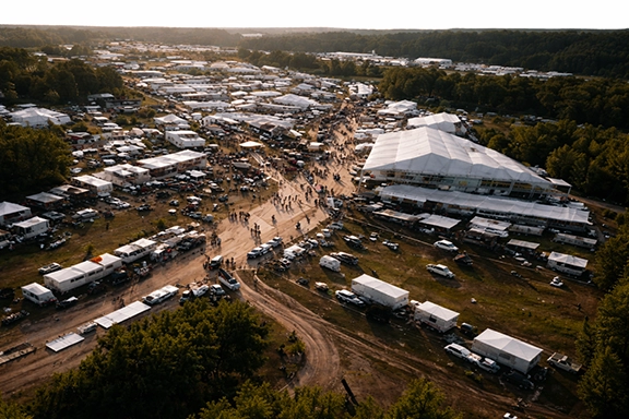 Aerial view of a large outdoor event with multiple white tents, vehicles, and people walking on dirt roads surrounded by trees.