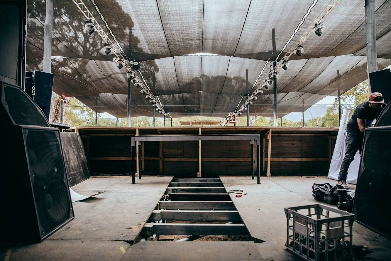 Empty outdoor stage under mesh canopy with speakers, tools, and a person working on the right side.