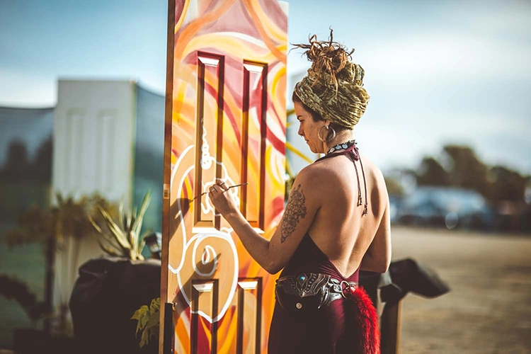 Woman with a headwrap painting colorful swirls on a standing door outdoors in bright sunlight.