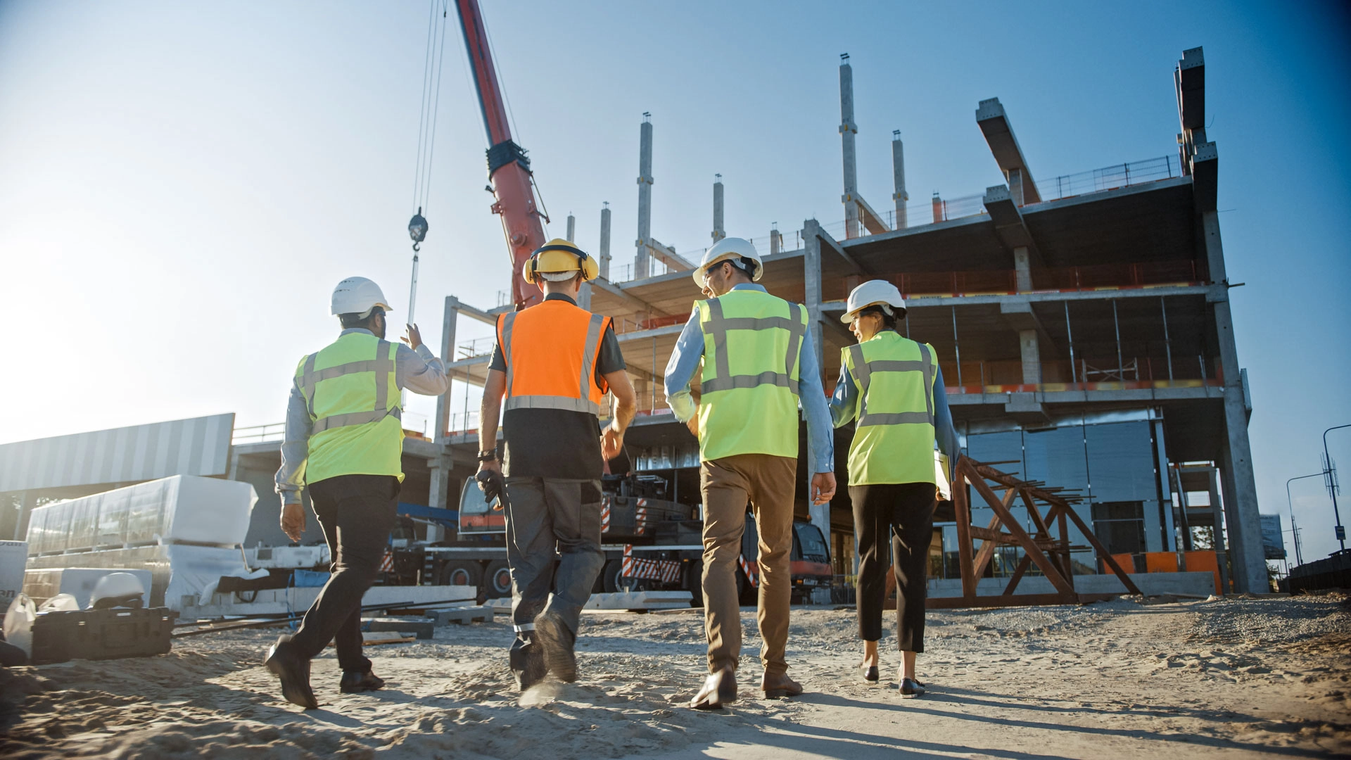 Four construction workers in safety vests and helmets walking toward a partially built multi-story building under a clear blue sky.