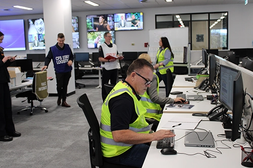 Office workers wearing high-visibility vests collaborate and work at computers in a modern control room.
