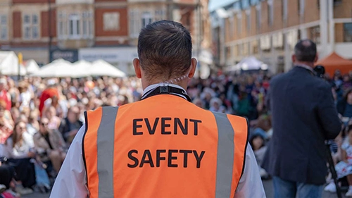 Person wearing an orange vest labeled 'EVENT SAFETY' overseeing a crowded outdoor event.