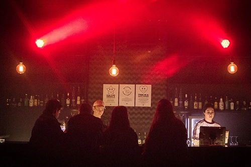 Dimly lit bar scene with four patrons seated and two bartenders illuminated by hanging bulbs and red stage lights.