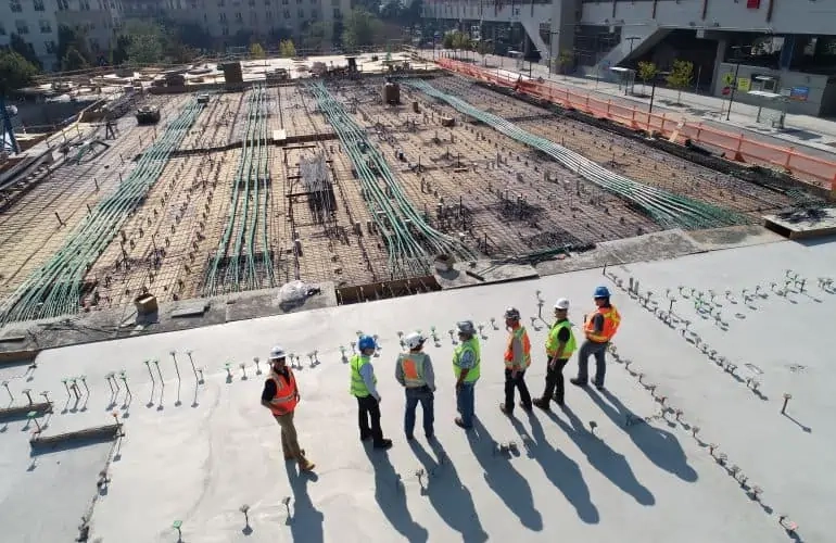 Seven construction workers in safety vests and helmets standing on a concrete surface overlooking a large construction site with exposed rebar and green conduits.