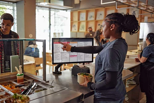 Restaurant worker pointing at a screen behind a counter while holding a bowl of food and interacting with a customer.