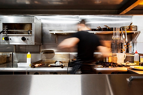 Blurred chef cooking in a busy commercial kitchen with stainless steel appliances and utensils.
