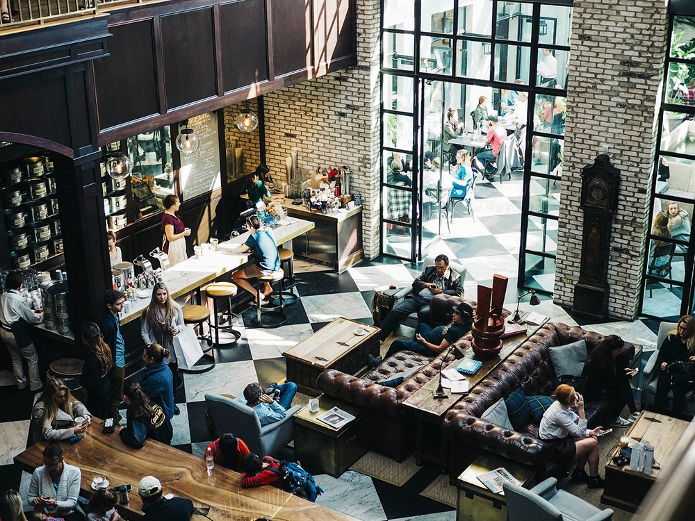Busy cafe interior with people sitting on leather sofas, chairs, and stools, enjoying drinks and using phones near large windows.