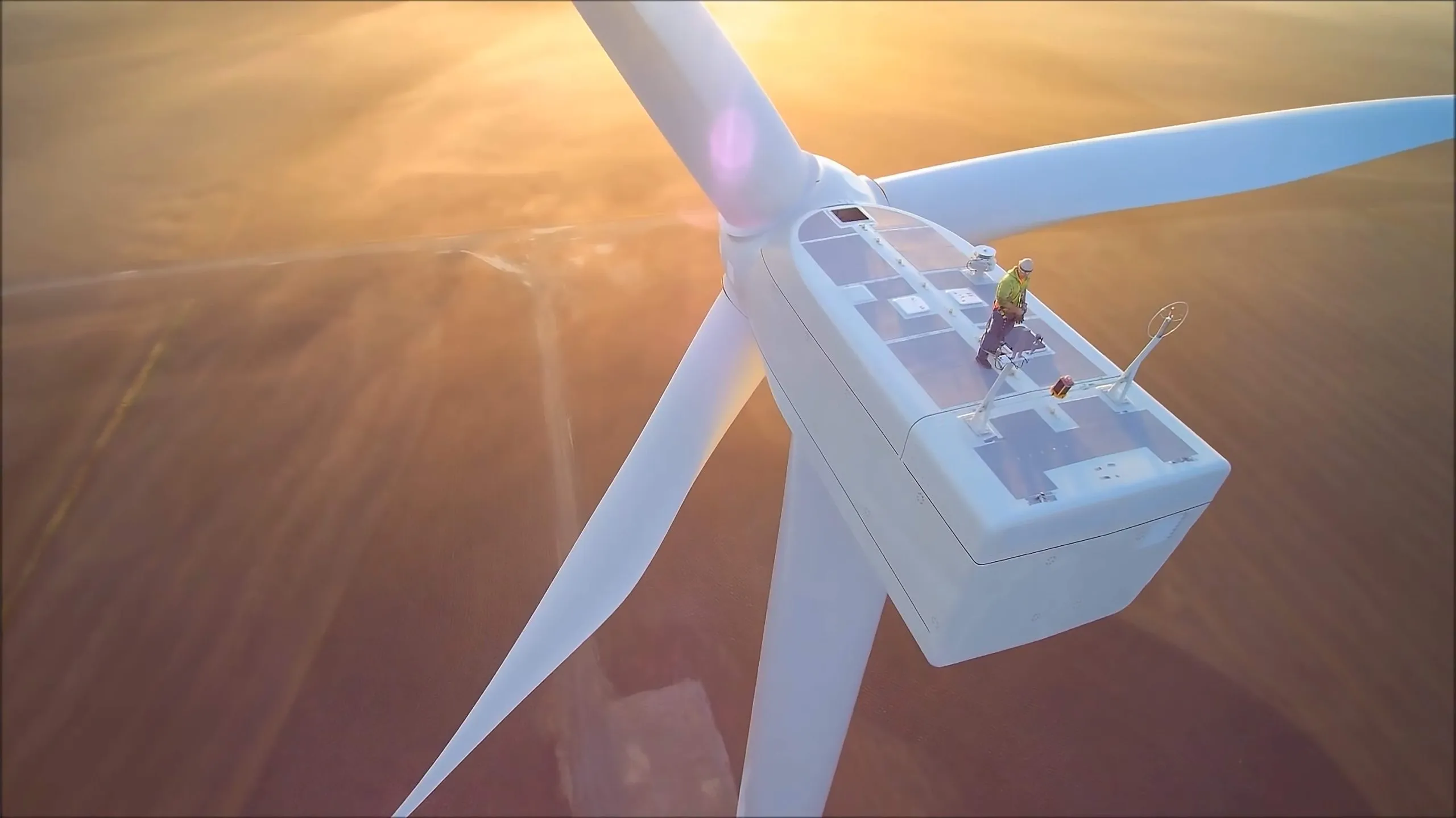 Technician standing on top of a large wind turbine at sunset with the blades extended over farmland.