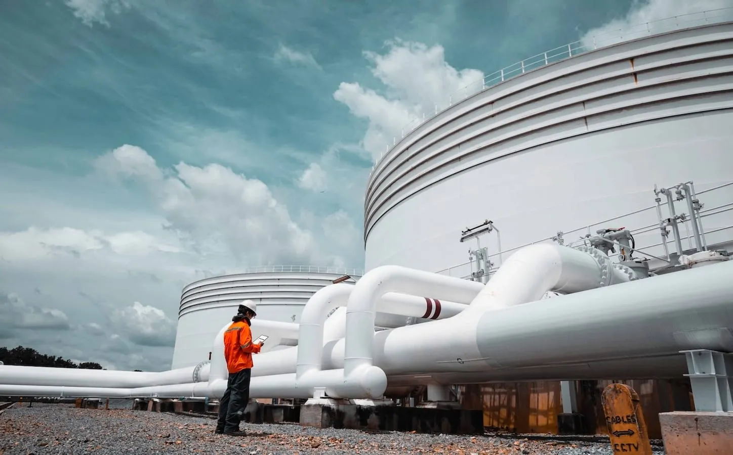 Worker in orange safety jacket and helmet inspecting large white industrial pipes near storage tanks under a cloudy sky.