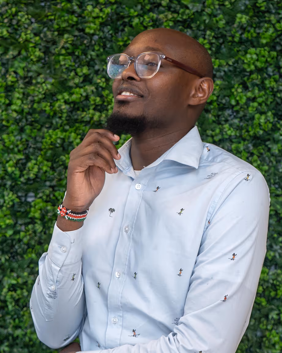 Man wearing glasses and a light blue shirt with small colorful figures, standing thoughtfully in front of a green leafy background.