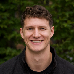 Young man with short curly hair smiling outdoors with green foliage background.