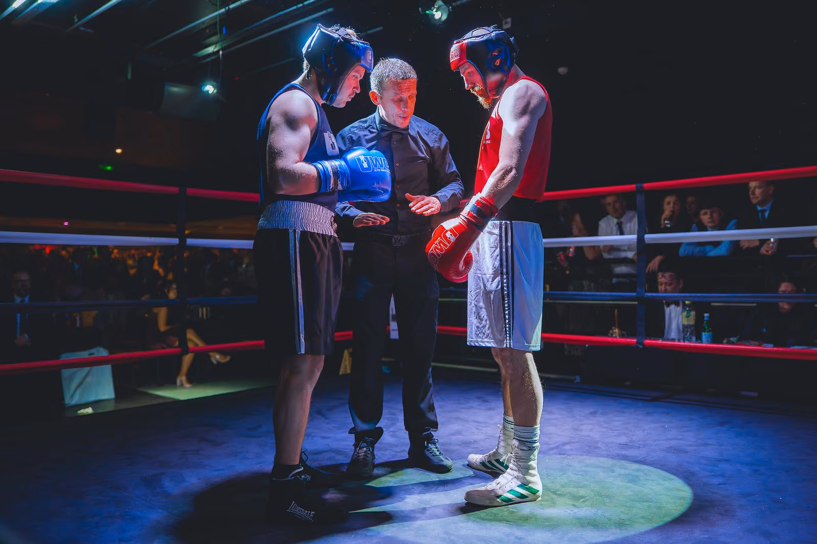 Two boxers wearing headgear and gloves listening to a referee inside a boxing ring under spotlight.
