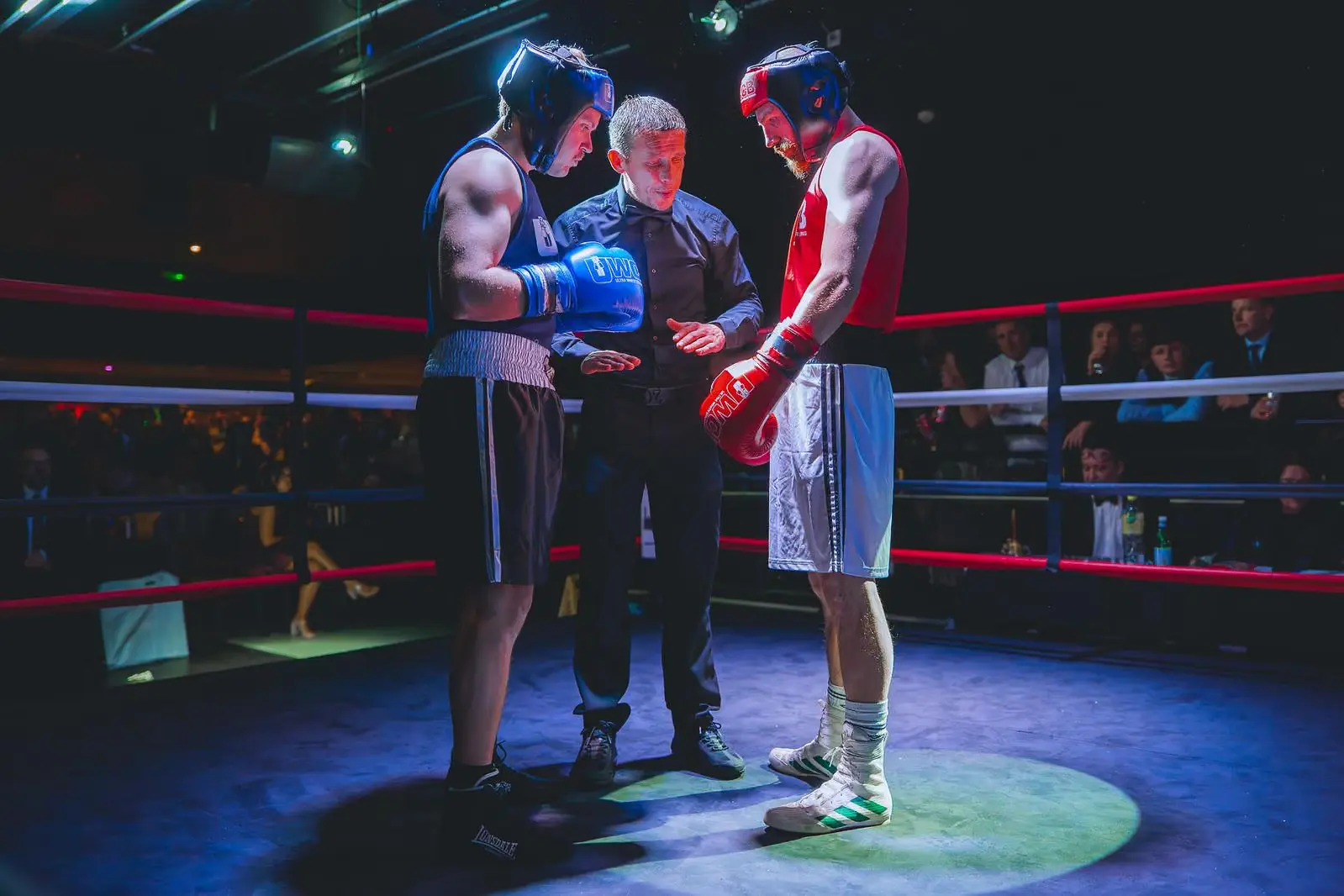 Two boxers wearing headgear and gloves listening to a referee inside a boxing ring under spotlight.