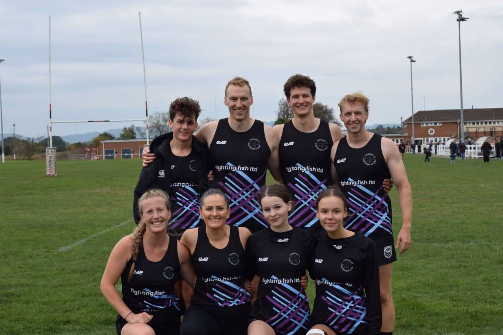 Group of eight rugby players in black and purple team uniforms posing on a grassy field with rugby goalposts and buildings in the background.