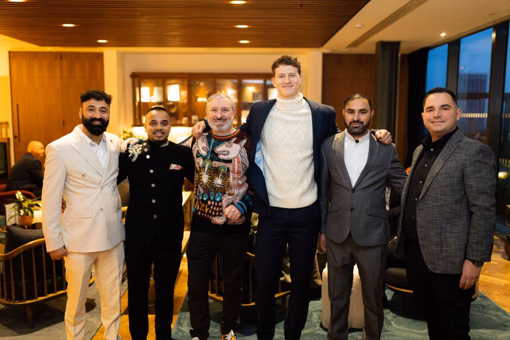 Six men standing indoors posing for a group photo, dressed in various formal and semi-formal outfits.