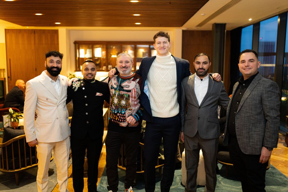 Six men standing indoors posing for a group photo, dressed in various formal and semi-formal outfits.