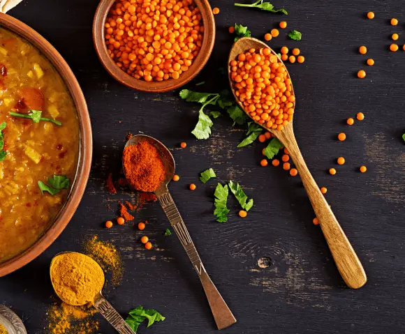 Wooden bowls and spoons with red lentils, turmeric powder, and red chili powder on a dark wooden surface, alongside a bowl of lentil soup garnished with herbs.