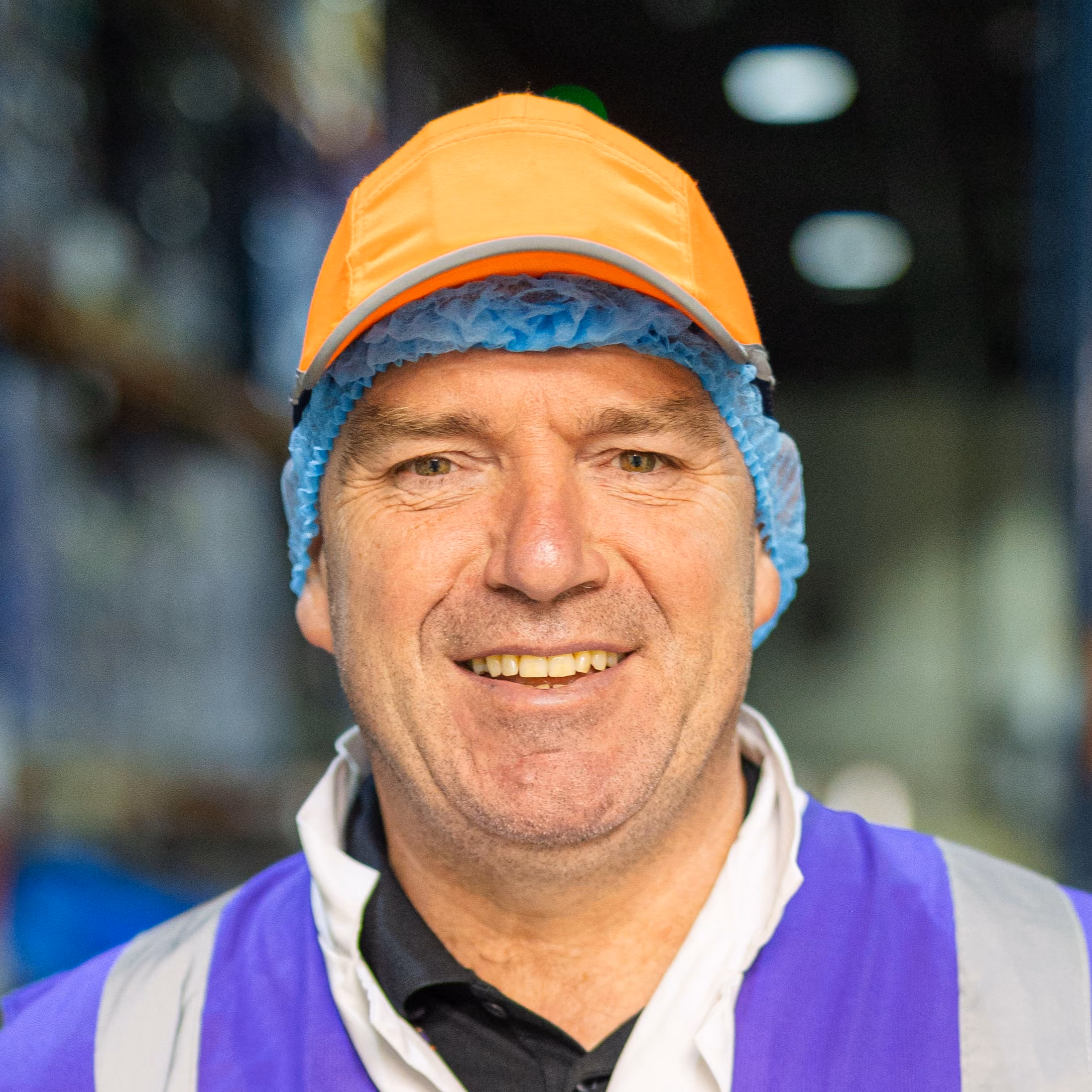 Smiling middle-aged male worker wearing an orange cap and blue hairnet in a warehouse.