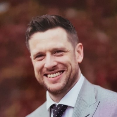 Smiling man in a gray suit jacket and white shirt with a navy blue polka dot tie.