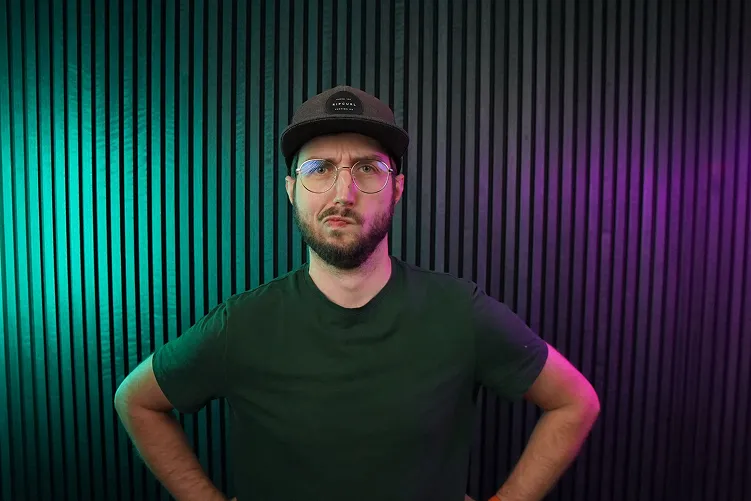 Man with beard, glasses, and black cap standing with hands on hips against a vertical black-striped background lit with green and purple lights.
