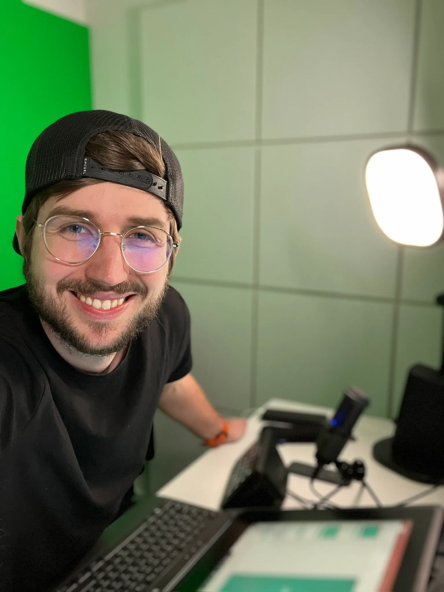 Nico Thümler with glasses and a backward black cap leaning over a keyboard with a microphone and ring light in the background.