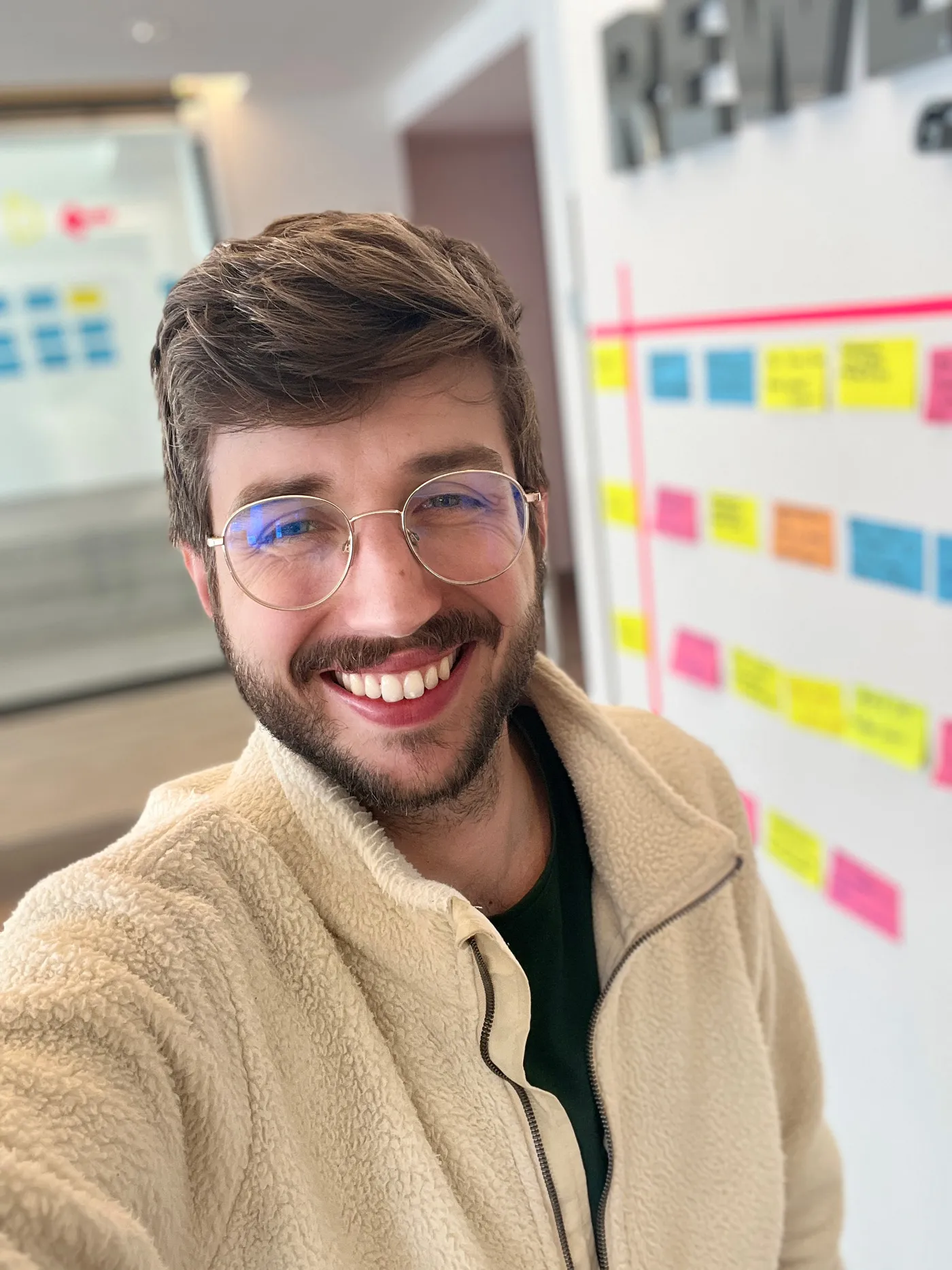 Nico Thümler smiling with glasses and brown hair wearing a beige fleece jacket indoors, with a blurred whiteboard covered in colorful sticky notes in the background.