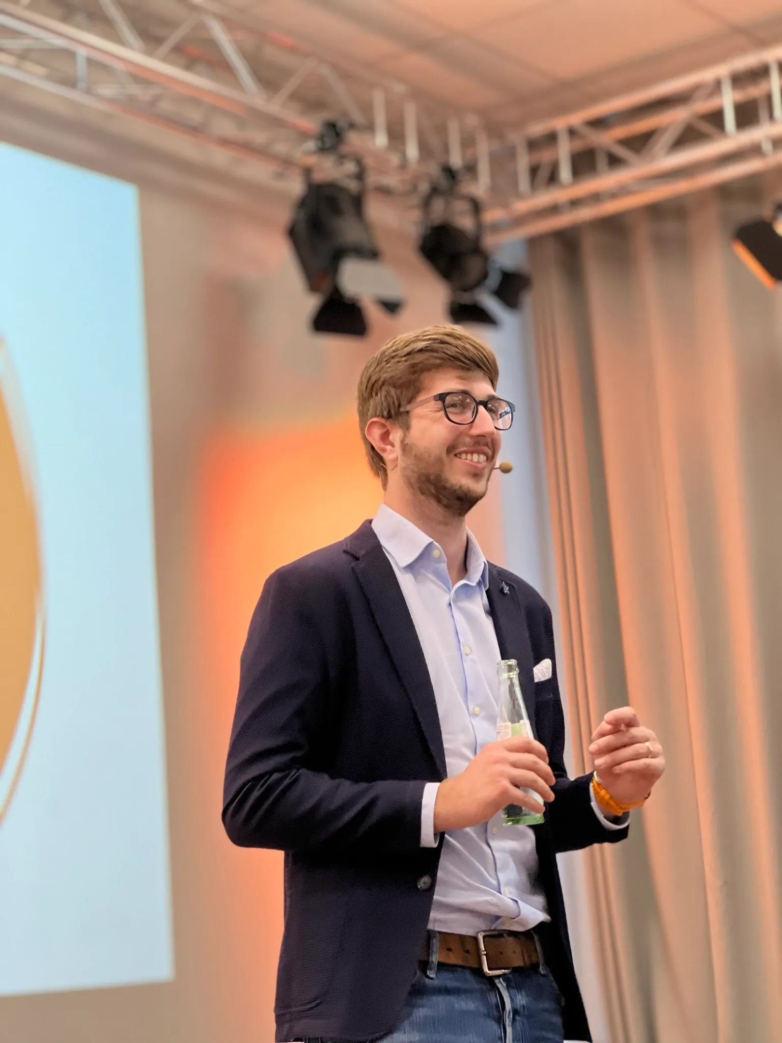 Nico Thümler wearing glasses, a blazer, and jeans, holding a bottle and speaking on stage with a headset microphone.