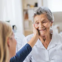 Jüngere Frau berührt liebevoll das Gesicht einer älteren lächelnden Frau mit grauen Haaren.