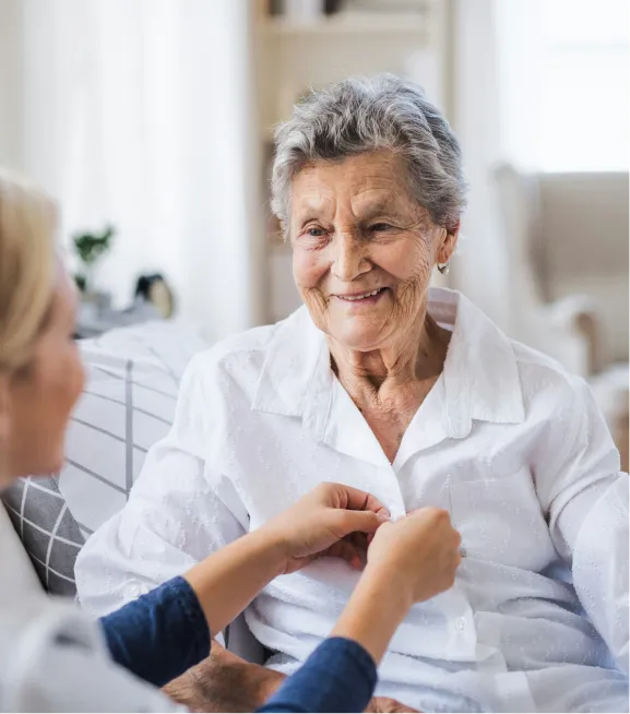 Eine ältere Frau lächelt, während eine jüngere Person ihre Bluse zumacht.