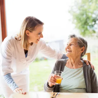 Pflegekraft lächelnd mit älterer Frau, die ein Glas Saft hält.