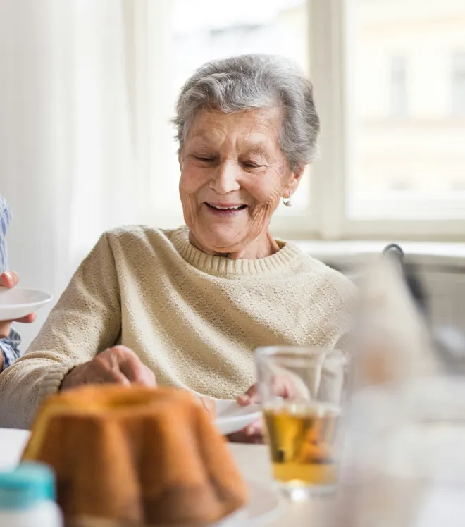Eine lächelnde ältere Frau in einem beigefarbenen Pullover sitzt am Tisch mit Gebäck und einem Glas Getränk.