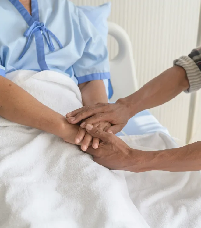 Jemand hält die Hände eines im Krankenhausbett liegenden Patienten im hellblauen Krankenhaushemd.