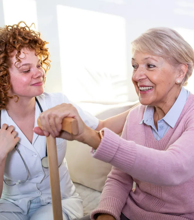 Pflegekraft mit Stethoskop hält freundschaftlich die Hand einer älteren Frau, die einen Stock hält.