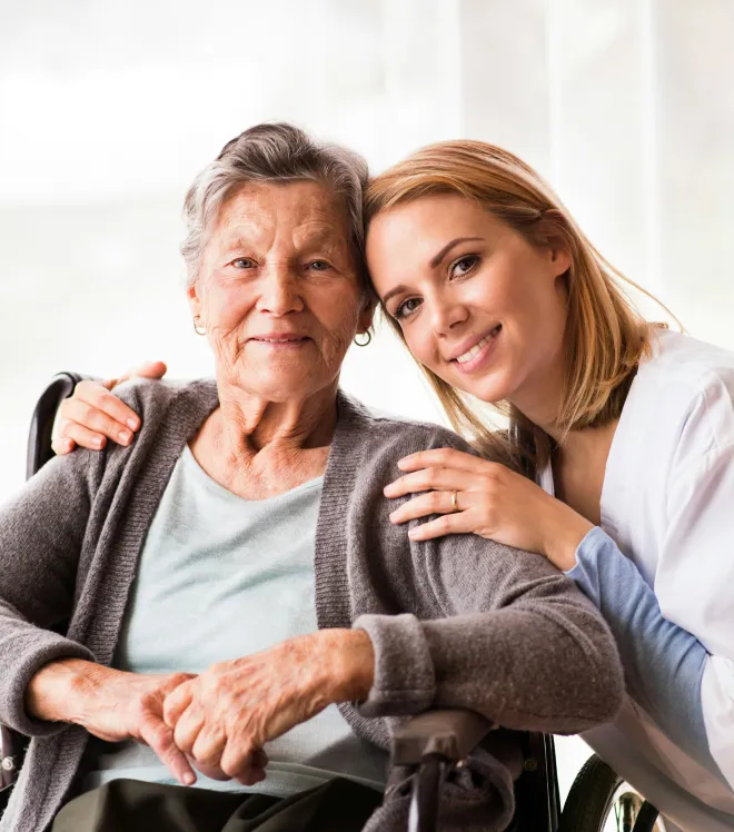 Ältere Frau im Rollstuhl mit jüngerer Frau, die sie freundlich umarmt.