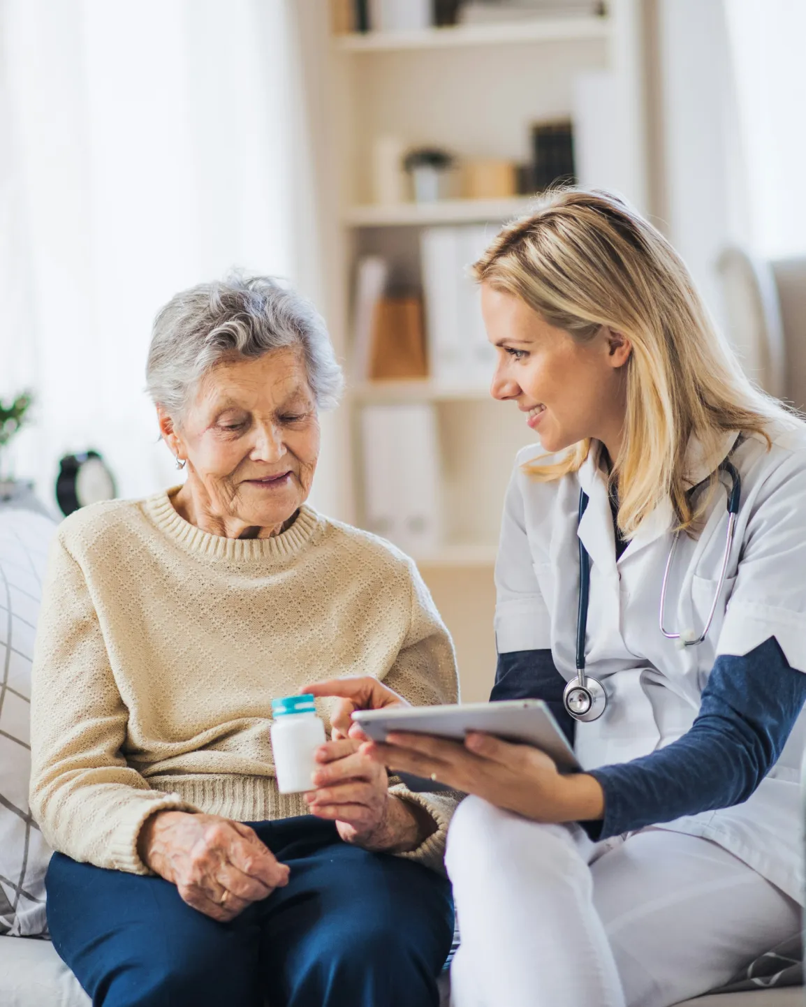 Ältere Frau hält Medikamentenflasche, während eine Krankenschwester mit Stethoskop sie lächelnd berät.