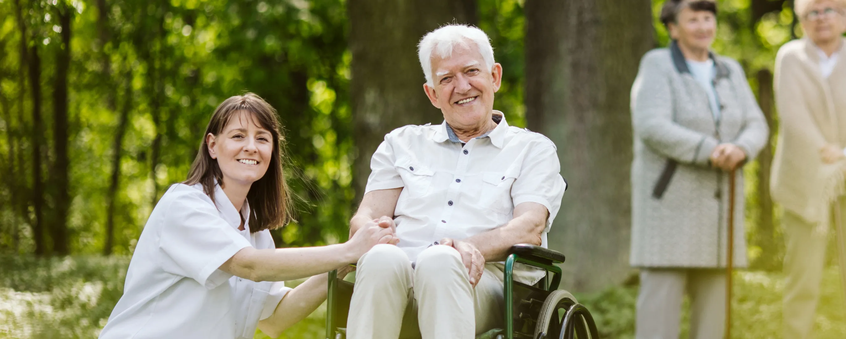 Pflegekraft hält die Hand eines lächelnden älteren Mannes im Rollstuhl in einem Park.