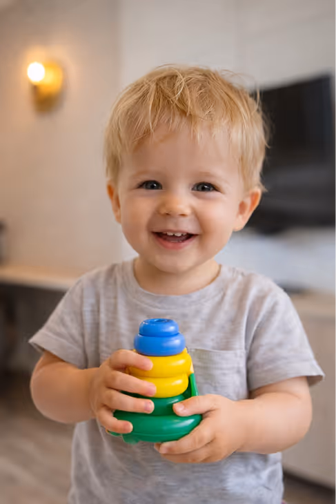 Smiling toddler with blonde hair holding a colorful plastic stacking toy in a cozy room.