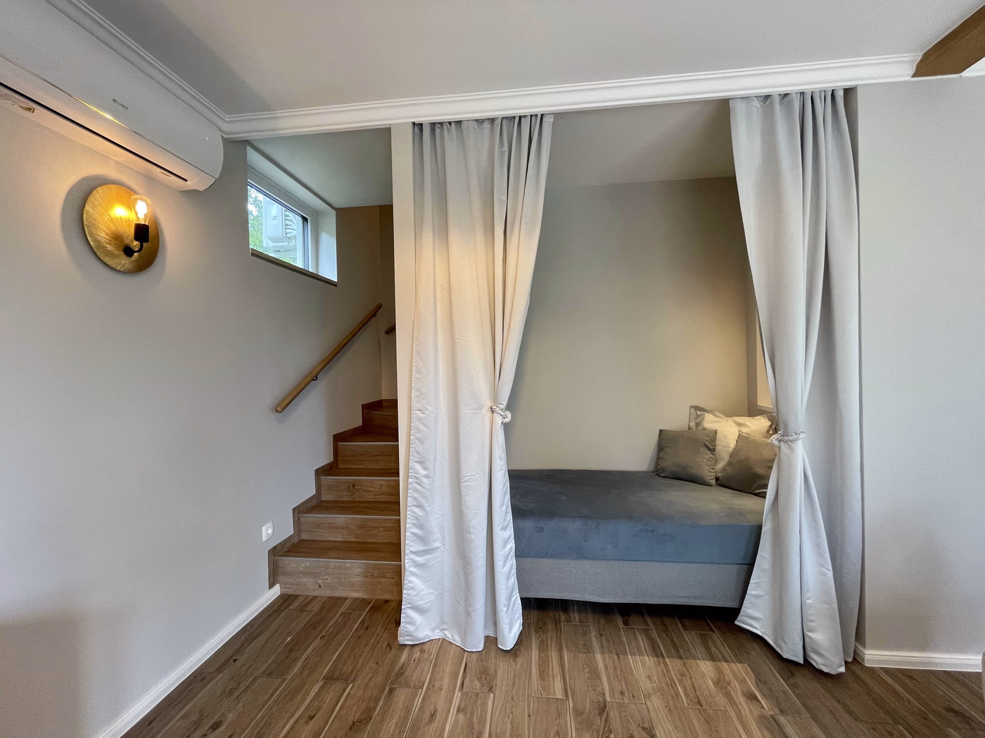 Room with wooden stairs on the left and a bed with gray cushions behind tied-back white curtains on the right.