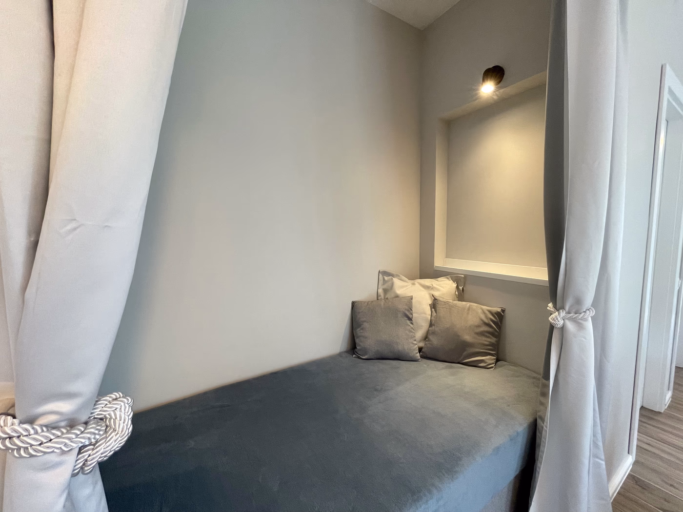 Cozy small nook with a padded bench, three pillows, gray curtains tied back, and a wall light above.