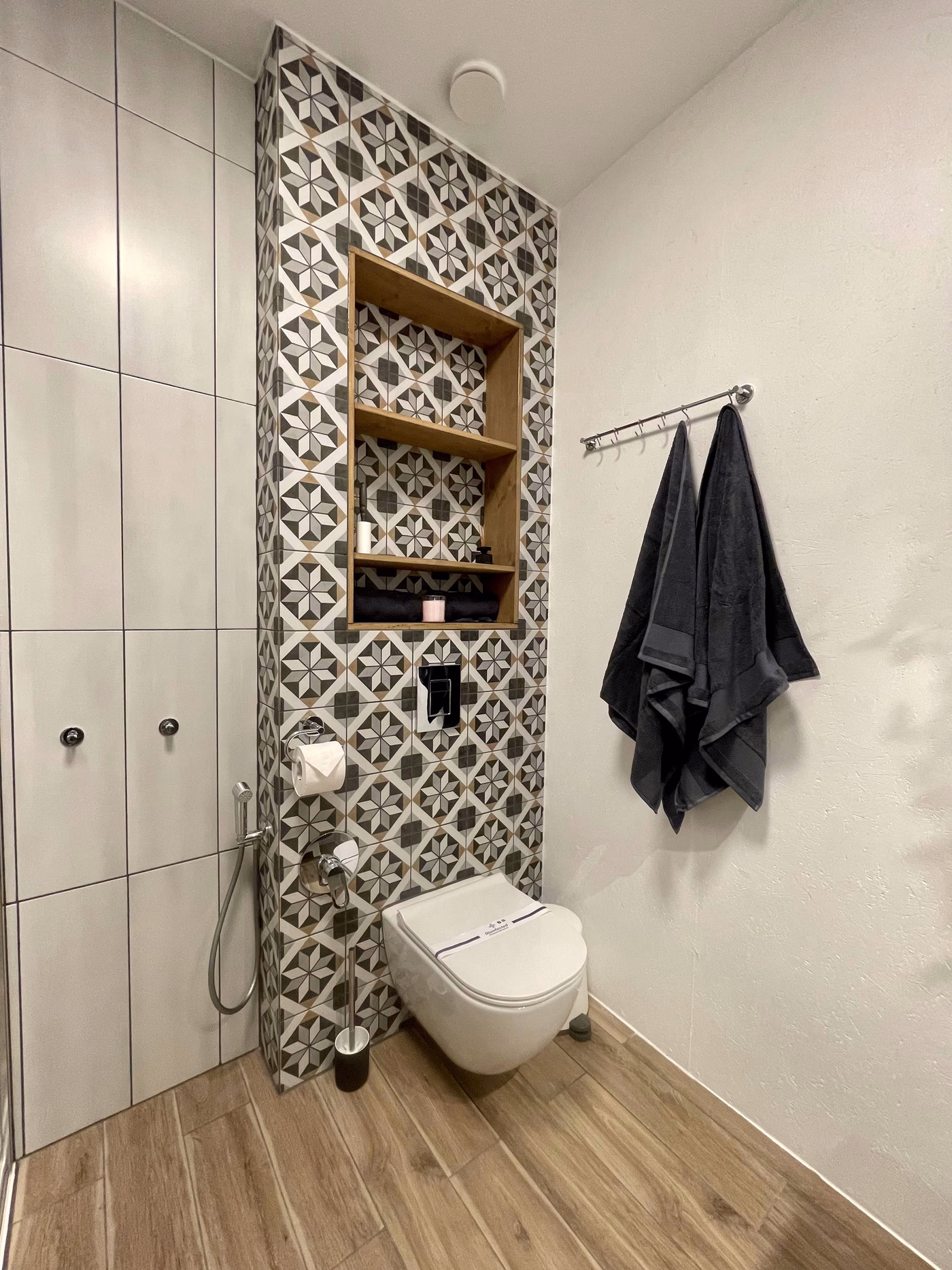 Modern bathroom corner with a wall-mounted toilet, patterned tile wall with built-in wooden shelves, and two dark towels hanging on a metal rack.