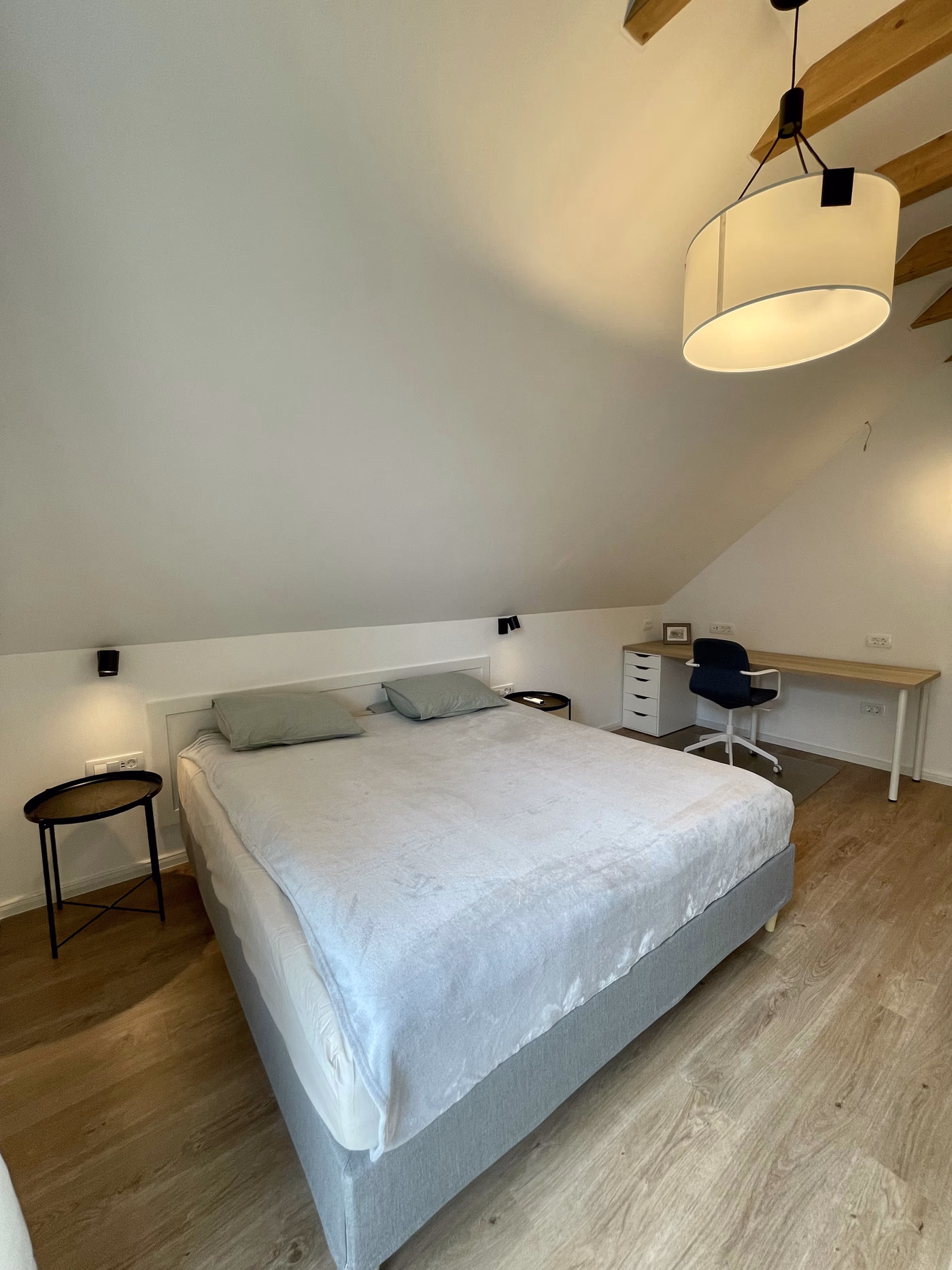Minimalist bedroom with a large bed covered in a light gray blanket, two pillows, wooden floor, a small round side table, a study desk with a swivel chair, and a ceiling lamp with wooden beams above.