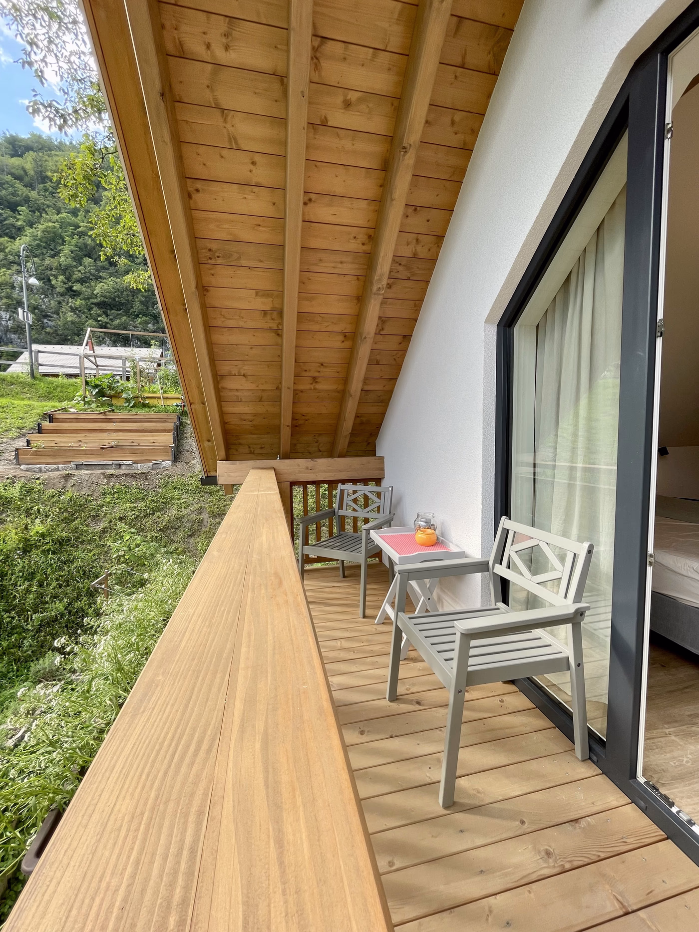 Wooden balcony with two gray chairs and a small table holding an orange candle jar, overlooking a grassy hill and garden beds.