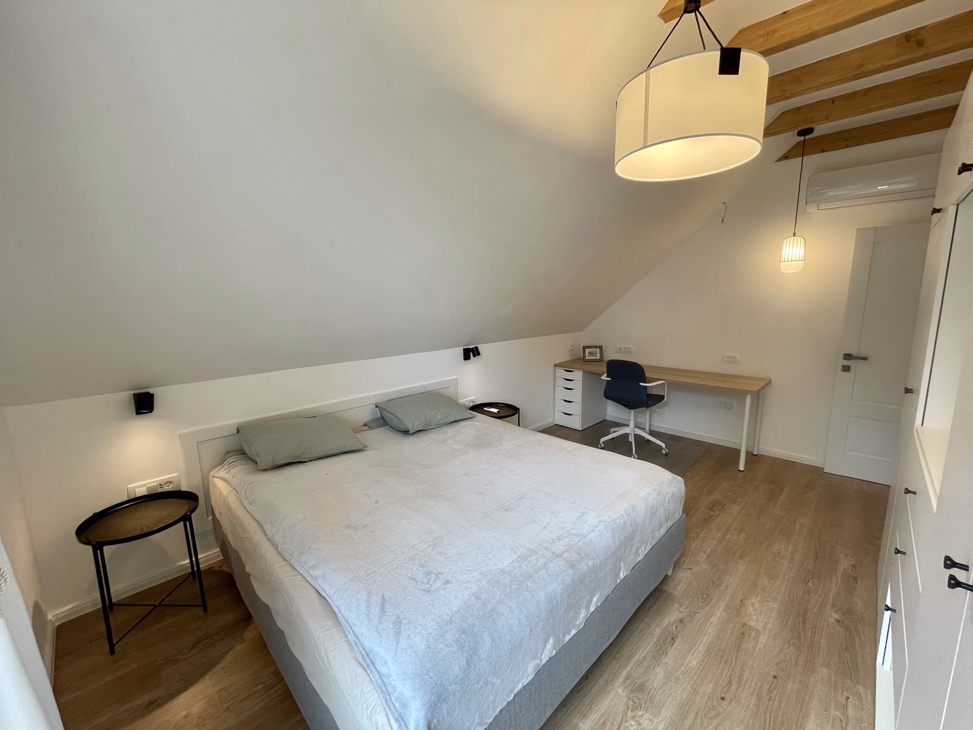 Minimalist bedroom with a large bed, light grey bedding, two green pillows, wooden floor, a desk with a chair, and ceiling lights.