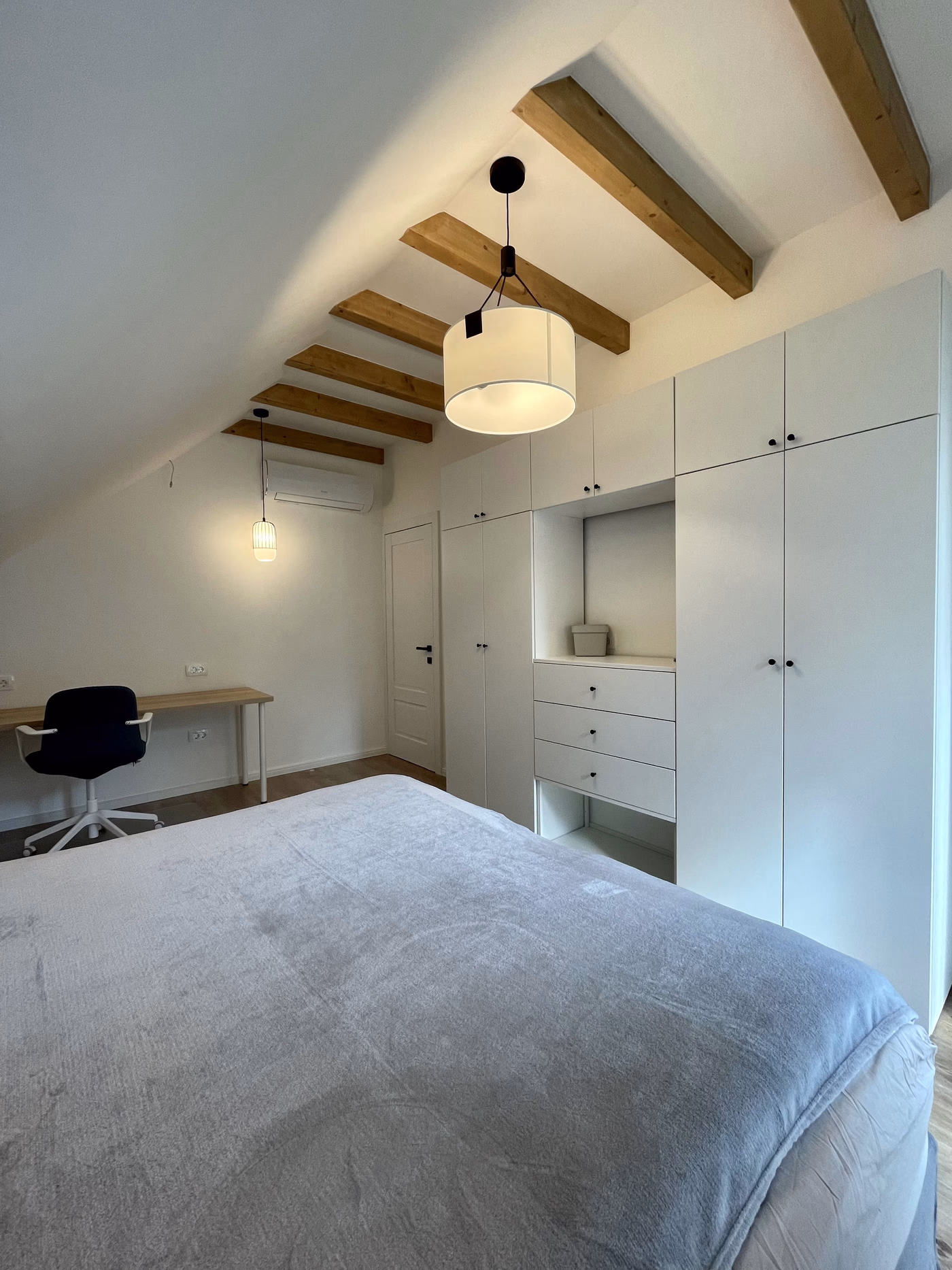 Minimalist bedroom with wooden ceiling beams, white built-in wardrobes, a grey bedspread, a desk, and two hanging pendant lights.